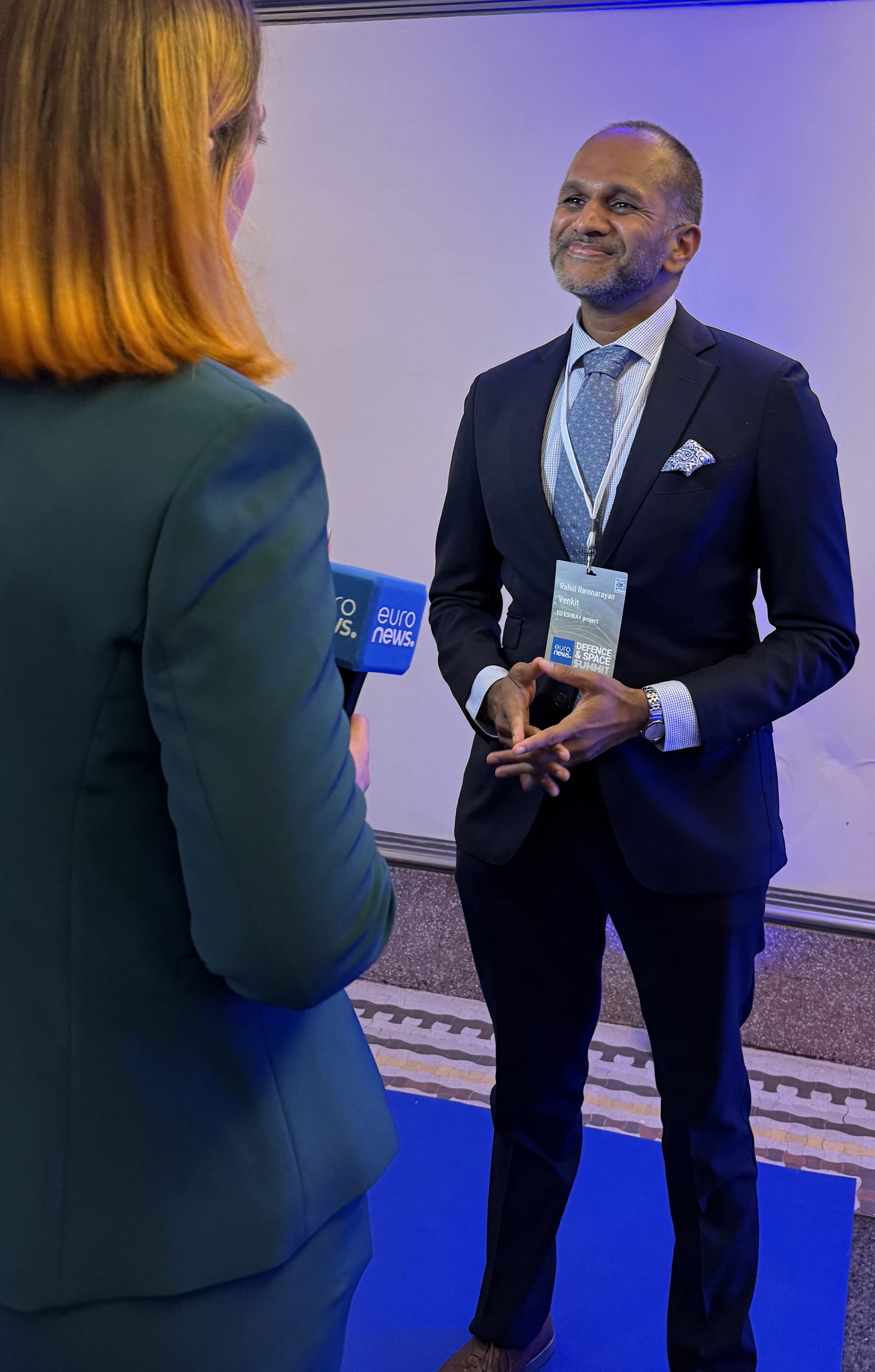 Man in a dark suit with a conference lanyard, hands clasped